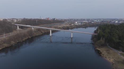 A-drone-approaches-a-long-bridge-over-a-river,-capturing-its-span-and-the-surrounding-cityscape