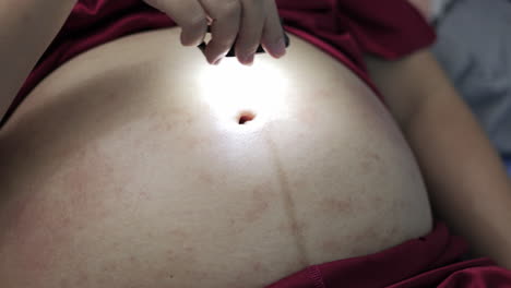 An-expectant-mother-is-holding-a-tiny-flashlight-and-moving-it-around-her-swollen-belly,-showing-stretched-marks-and-some-scratches-on-her-skin