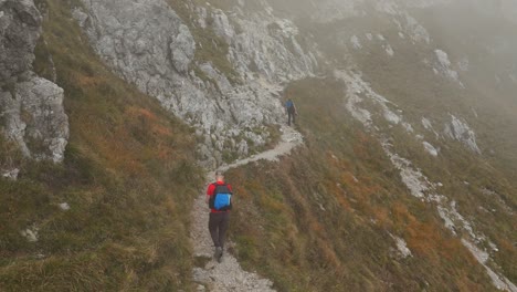 Back-view-of-two-hikers-walking-along-path-of-Resegone-mountain-in-northern-Italy-with-fog-in-background-on-misty-day