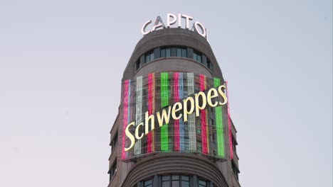 A-vibrant-Schweppes-colorful-neon-sign-adorns-the-Carrion-building,-known-as-the-Capitol,-on-Madrid's-Gran-Via,-a-bustling-hub-for-evening-entertainment-and-retail