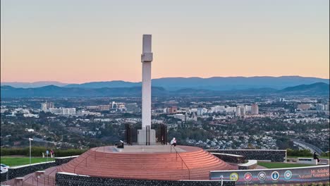 Gegen-Den-Uhrzeigersinn-Verlaufende-Kreisförmige-Drohnen-Hyperlapse-Des-Mount-Soledad-Gedenkkreuzes,-Wobei-Die-Sonne-Beim-Zweiten-Durchgang-Sichtbar-Ist-Und-Sich-Die-Sonne-Mit-Den-Lücken-Im-Kreuz-überschneidet,-Während-Sich-Menschen-Versammeln