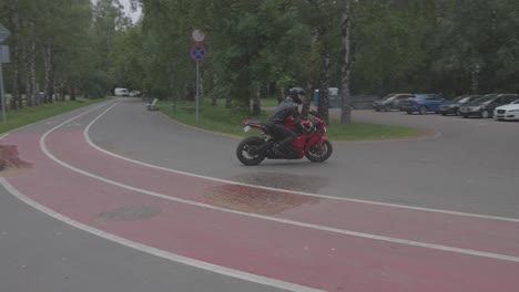 4k,-Eine-Person-In-Einem-Roten-Motorrad-Und-Einem-Schwarzen-Anzug-Fährt-Die-Straße-In-Einem-Park-Entlang,-Im-Hintergrund-Ein-See