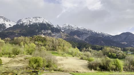 View-from-train-window-passing-majestic-snow-capped-mountains-towering-over-lush-green-valleys