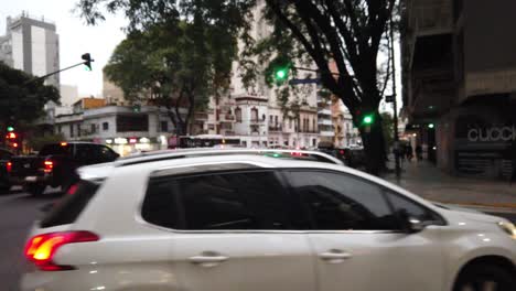 Los-Autos-Giran-A-La-Derecha-En-La-Calle-En-La-Avenida-Alberdi,-El-Tráfico-Del-Horizonte-Al-Anochecer-En-La-Ciudad-De-Buenos-Aires,-Argentina,-Peatones-Y-Edificios-Del-Barrio-De-Flores.