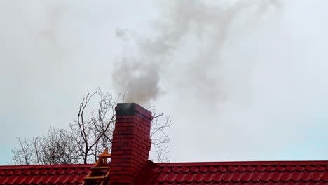 Thick-smoke-spew-through-house-chimney,-cherry-red-roof-contrasting-with-grey-gloomy-sky