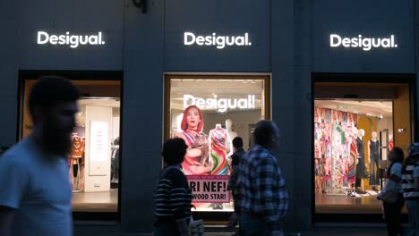 Shoppers-and-pedestrians-walk-past-the-Spanish-clothing-brand-Desigual-store-at-nighttime