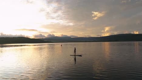 Frau-Steht-Bei-Tagesanbruch-Auf-Einem-Paddleboard-Auf-Einem-See,-In-Der-Ferne-Ist-Die-Silhouette-Eines-Kajakfahrers-Zu-Sehen