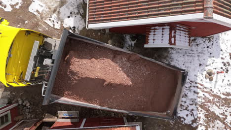 Tipper-truck-dumps-load-of-gravel-next-to-red-building-in-winter,-drone-overhead