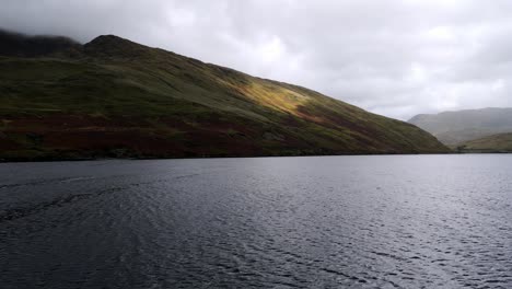 Drone-flight-over-a-large-lake-or-loch-in-the-Scottish-highlands-with-green-rolling-mountains-and-a-road-that-follows-the-shoreline