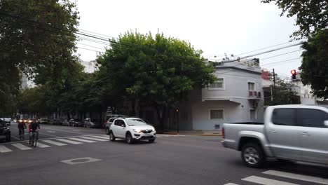 Cars-driving-through-avellaneda-avenue-flores-neighborhood-traffic-at-buenos-aires-city-dusk-skyline-trees-autumn,-pedestrians-and-stores