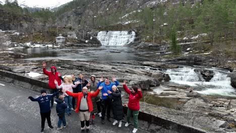 Happy-tourist-group-waving-and-smiling-in-front-of-springtime-waterfall-during-guided-tour