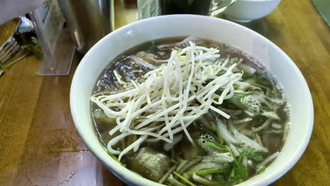 Sopa-De-Fideos-Vietnamita-Fresca-Sobre-Una-Mesa-En-Un-Tazón-Añadiendo-Ingredientes,-Toma-Manual