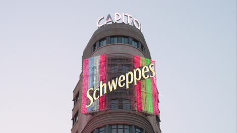 Eine-Leuchtende-Und-Farbenfrohe-Neonreklame-Der-Schweizer-Getränkemarke-Schweppes-Ist-An-Der-Fassade-Des-Aasgebäudes-Der-Gran-Via-Zu-Sehen,-Das-Als-Capitol-Bekannt-Ist