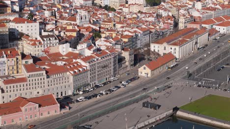 Aerial-tracking-along-roadway-by-Tagus-River-with-cars-driving-at-midday-below-orange-cathedral-and-home-roofs-in-Lisbon-Portugal