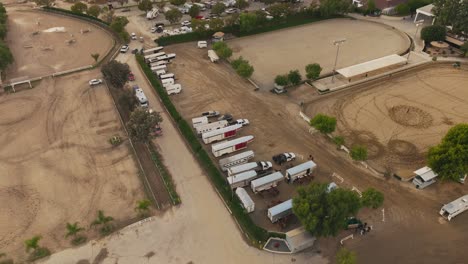 Toma-Aérea-Del-Centro-Ecuestre,-Remolques-Y-Camiones-Moviéndose-Lentamente-Por-El-Lote,-Patios-De-Librea-Con-Caballos-Y-Entrenadores-Debajo