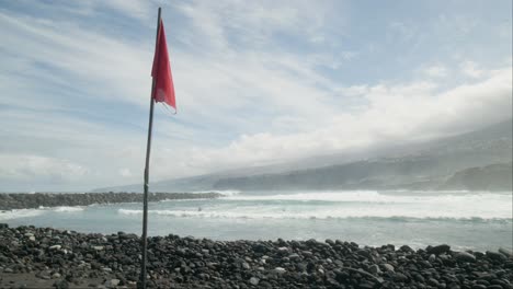 Red-warning-flag-with-slowmotion-group-of-surfers-surfing-on-ocean-waves-on-pebble-beach,-Playa-Martiánez,-Puerto-de-la-Cruz,-Tenerife,-Canary-Islands-in-spring