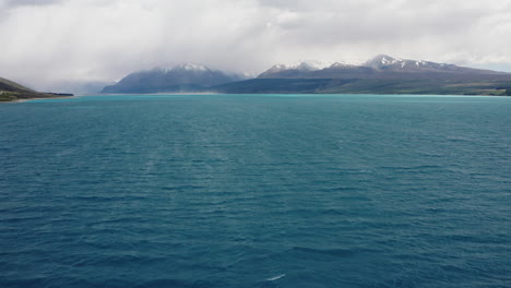 Fly-over-bright-blue-water-on-Lake-Pukaki-while-low-clouds-shroud-the-surrounding-snow-capped-picturesque-mountains
