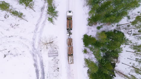 Truck-loaded-with-harvested-tree-logs-drive-on-snowy-forest-road,-drone-overhead