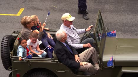 Veteran,-his-wife-and-grandchildren-riding-on-old-vintage-military-vehicle-down-the-street-of-Brisbane-city,-participating-in-annual-Anzac-Day-parade,-waving-at-the-cheering-crowds,-close-up-shot
