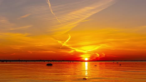 Vibrant-timelapse-sunset-over-Baltic-Sea-in-Tallinn,-Estonia,-casting-a-warm-glow-on-the-water-and-the-city-skyline,-with-silhouettes-of-anchored-boats,-as-streaks-of-clouds-drift-across