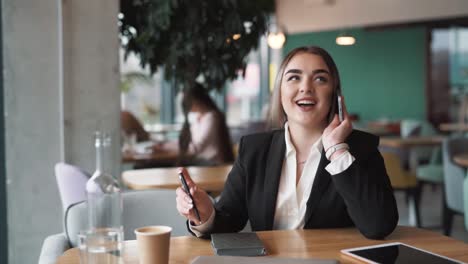 Portrait-of-a-young,-beautiful-woman-sitting-in-a-café,-dressed-in-business-attire,-talking-on-the-smartphone-with-a-smile-on-her-face