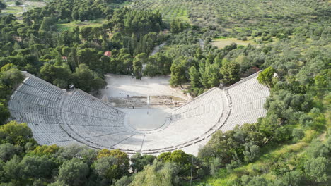 Aerial-drone-shot-of-the-Epidaurus-Theatre-in-Greece,-surrounded-by-lush-greenery-with-a-single-person-standing-in-it,-highlighting-the-vastness-of-the-ancient-structure