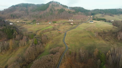 A-winding-road-cuts-through-rolling-hills-and-sparse-woodlands,-with-a-hint-of-rural-habitation-in-the-backdrop