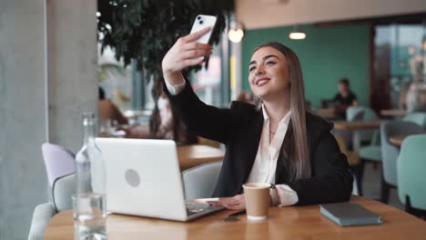 Hermosa-Mujer-Sentada-En-Un-Café-Con-Traje-De-Negocios-Se-Toma-Una-Selfie-Con-Una-Sonrisa-En-Su-Rostro-En-Un-Teléfono-Inteligente