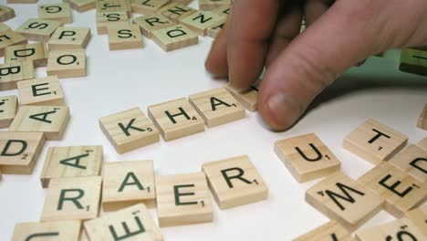Male-hand-with-Scrabble-tile-letters-makes-word-KHAT-on-table-top