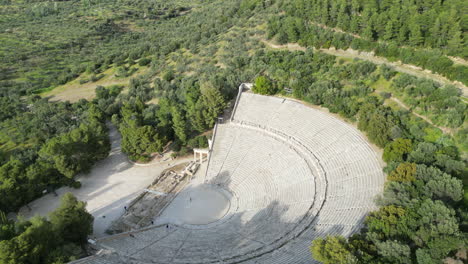 Aerial-drone-shot-of-the-Epidaurus-Theatre-in-Greece,-showcasing-its-grand-scale-of-theatre-compared-to-a-person-at-its-center,-surrounded-by-trees