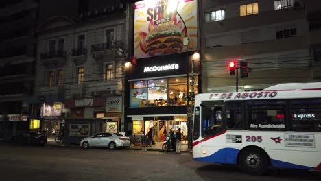 Tienda-De-Comida-Mc-Donald&#39;s-Por-La-Noche-En-La-Avenida-De-La-Ciudad-De-Buenos-Aires-Recorrido-En-Autobús-Por-La-Escena-Nocturna-Paisaje-Urbano-Del-Barrio-Caballito-La-Gente-Camina,-Argentina