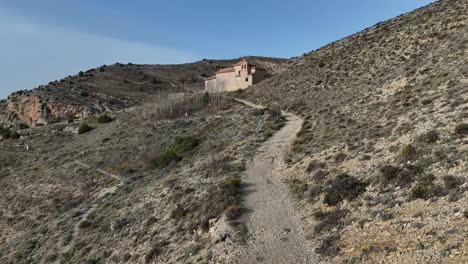 Hermitage-approaching-drone-view-following-a-path-in-an-arid-desolate-landscape-in-Albarracin-Village,-Teruel,-Spain