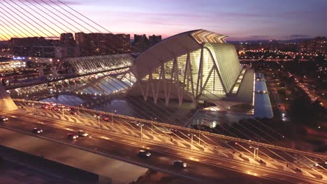 Puesta-De-Sol-Y-Luces-De-La-Ciudad,-Sobre-La-&quot;ciudad-De-Las-Artes-Y-Las-Ciencias&quot;-De-Valencia:-Una-Vista-Cinematográfica,-El-Tráfico-Cruzando-El-Puente-En-La-Hora-Dorada