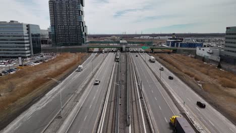 Vista-Aérea-Frontal-Del-Sistema-Automatizado-De-Tren-Ligero-Réseau-Express-Métropolitain-Rem-En-Brossard,-Cerca-De-La-Ciudad-De-Montreal,-Conduciendo-Por-La-Carretera-Principal-Con-Tráfico-De-Automóviles