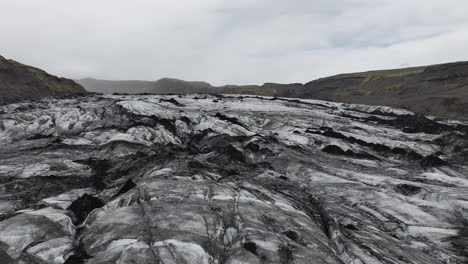 Volando-Sobre-Un-Glaciar,-Una-Capa-De-Hielo-Blanca-Y-Negra-Bajo-Colinas-Volcánicas,-Islandia