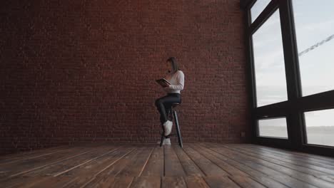 Mujer-Sentada-En-Un-Asiento-Alto-En-Una-Habitación-Vacía-Cerca-De-Una-Ventana-Brillante-Y-Una-Pared-De-Ladrillo-Rojo