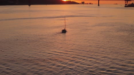 Aerial-establishing-dolly-tilt-up-follows-sailboat-on-Tagus-River-with-sunset-between-suspension-bridge