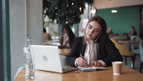 Mujer-Joven-Y-Hermosa-Sentada-En-Un-Café,-Vestida-Con-Traje-De-Negocios,-Hablando-Por-Teléfono,-Tomando-Notas-En-Su-Cuaderno-Y-Trabajando-En-Su-Computadora-Portátil.