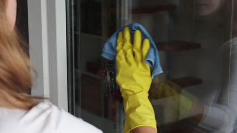 Woman,-housework-and-housekeeping-concept---woman-in-gloves-cleaning-window-with-rag-and-cleanser-spray-at-home