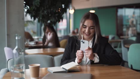 Portrait-of-a-young,-beautiful-woman-in-a-café,-wearing-business-attire,-receives-a-joyful-message-about-winning,-money,-and-victory