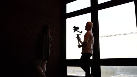 Videographer-with-camera-on-gimbal-film-model-walk-near-bright-studio-window