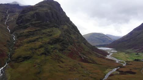 Dramatic-Aerial-backwards-shot-of-the-Glencoe-Mountains-Scotland-showing-a-river-flowing-down-the-mountain-side