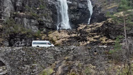 Minibus-passing-bridge-and-famous-waterfall-Hesjedalsfossen-in-slow-motion,-Aerial-Norway