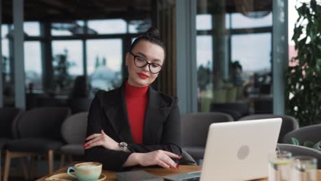 Eine-Wunderschöne-Junge-Frau-Mit-Brille-Und-Zahnspange,-Eine-Freiberuflerin,-Sitzt-Mit-Einem-Laptop-In-Einem-Stilvollen-Restaurant,-Trägt-Business-Kleidung-Und-Eine-Brille,-Lächelt-Und-Posiert-Für-Die-Kamera