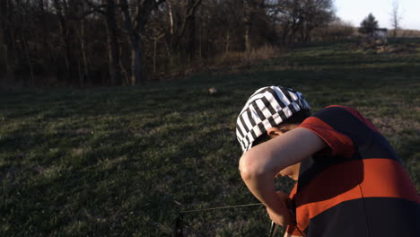 Un-Joven-Con-Gorra-A-Rayas-Practicando-Tiro-Con-Arco-En-Un-Campo-De-Hierba-Al-Atardecer,-Siloam-Springs,-Arkansas,-Vista-Lateral