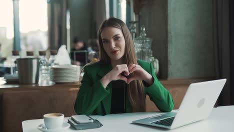 cheerful,-beautiful-young-woman-sits-in-a-stylish-restaurant,-speaking-while-looking-into-the-camera-with-a-smile,-showing-a-heart,-and-blowing-a-kiss