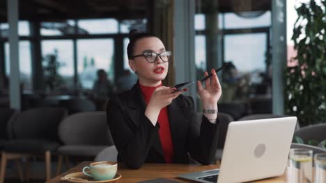 beautiful-young-woman-in-glasses-and-braces,-a-freelancer,-sits-in-a-stylish-restaurant-dressed-in-professional-attire,-recording-a-voice-message-on-her-smartphone-while-working-on-her-laptop