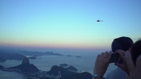 Tourists-marveling-at-the-vista-from-the-top-of-Corcovado-Mountain,-gazing-on-a-helicopter-,-down-upon-the-city-of-Rio-de-Janeiro-and-its-neighboring-isles-from-the-vantage-point-of-Cristo-Redentor