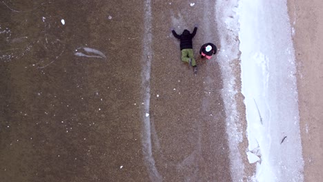 Woman-dragging-man-in-winter-clothing-by-leg-across-frozen-lake,-aerial