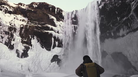 Turista-Caminando-En-Cámara-Lenta-Cerca-De-La-Cascada-De-Skogafoss-En-Temporada-De-Invierno,-Con-Paisaje-Escénico-De-Agua-Y-Hielo